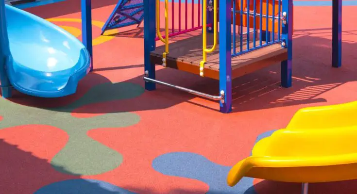 A playground with a blue slide and a yellow side, climbing equipment, and a rubber surface. The surface is red with blue designs.