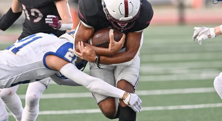 Two football teams playing against each other—a home player rushes with the ball as two opposing players attempt a tackle.