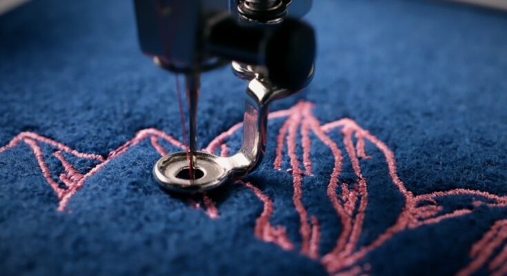 A close-up of an embroidery machine's needle and thread system stitching a pattern into blue fabric with pink thread.