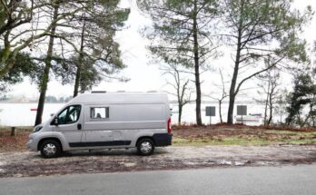 A white camper van parked on the side of a road with views of a waterfront. The shoreline has several pine trees.