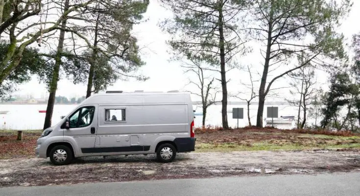 A white camper van parked on the side of a road with views of a waterfront. The shoreline has several pine trees.