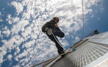 A firefighter using rope to rappel down the side of a fire station tower. The sky is blue and cloudy in the background.