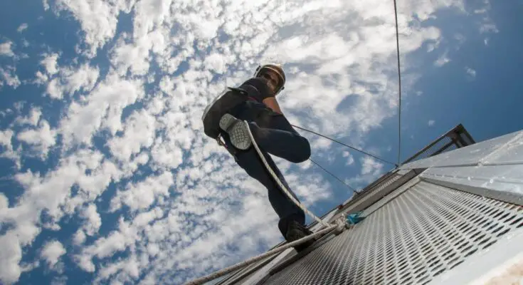 A firefighter using rope to rappel down the side of a fire station tower. The sky is blue and cloudy in the background.