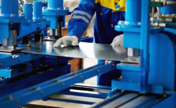 A worker dressed in a blue and yellow uniform is holding a piece of sheet metal in between a large blue machine.