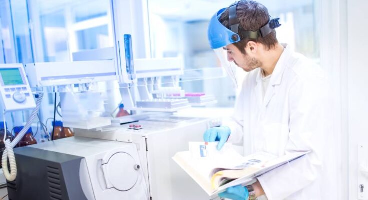 A scientist stands in a lab and holds a manual for a piece of equipment. He wears a shield over his face and blue gloves.