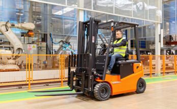 A man wearing a reflective safety vest is driving an orange and black forklift through a large warehouse.