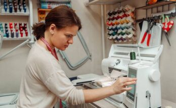 A woman in a white shirt with a pink tape measure around her neck. She's working on the screen of her embroidery machine.