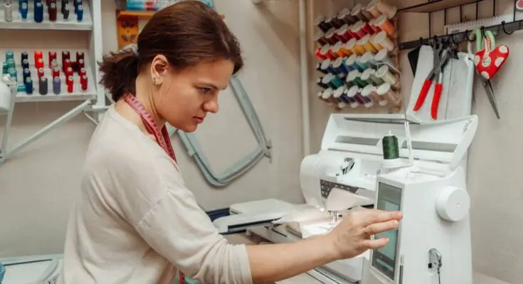 A woman in a white shirt with a pink tape measure around her neck. She's working on the screen of her embroidery machine.