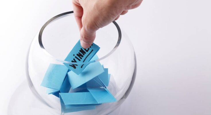 A hand is reaching into a glass bowl filled with blue slips of paper and pulling out a slip that says "winner" in black ink.