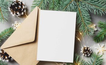 A close-up of a blank holiday card and envelope surrounded by pine tree branches, pine cones, and snowflake lights.