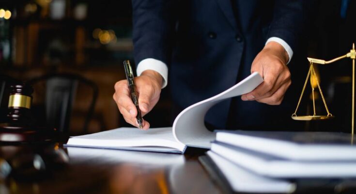 A lawyer in a dark-colored suit is opening a large notebook and using a fancy pen to write something in it.