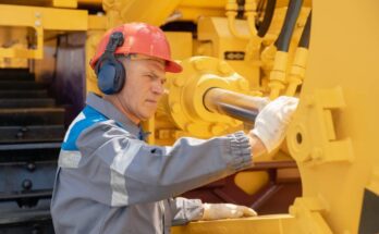 A man wearing a hard hat, hearing protection, and gloves inspects a component of a hydraulic system.