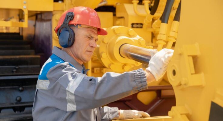 A man wearing a hard hat, hearing protection, and gloves inspects a component of a hydraulic system.