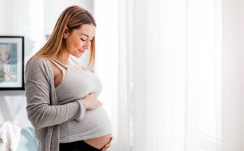 A pregnant woman in a gray tank top and cardigan standing near a sunny window, smiling as she looks down at her baby bump.