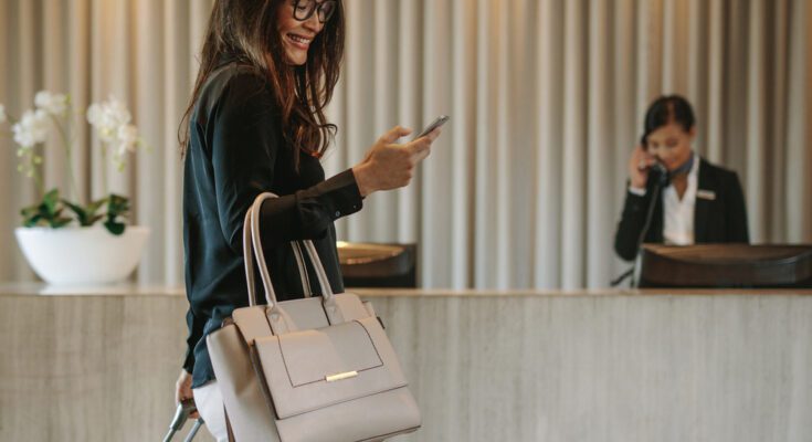 A woman smiles while looking at her phone. She walks through a hotel lobby while pulling a wheeled suitcase behind her.