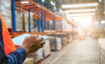 A close-up view shows a person holding a silver tablet while standing in the aisle of a warehouse.