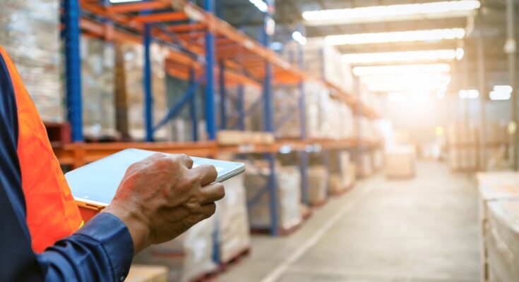 A close-up view shows a person holding a silver tablet while standing in the aisle of a warehouse.