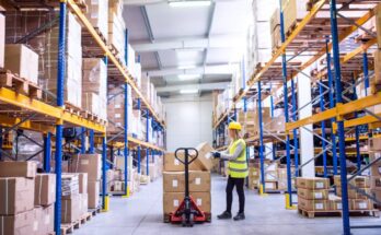 A woman in a bright yellow safety vest and hard hat places a cardboard box onto a stack of other boxes in a warehouse aisle.