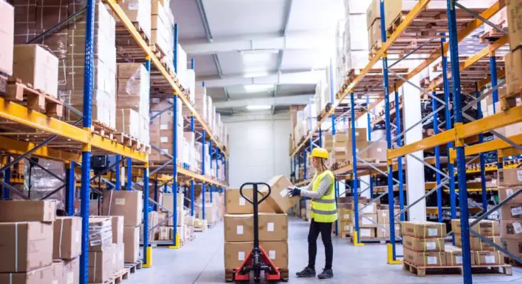 A woman in a bright yellow safety vest and hard hat places a cardboard box onto a stack of other boxes in a warehouse aisle.