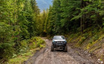 A silver-lifted truck drives on a dirt road that's covered in rocks and surrounded by tall, mature trees.