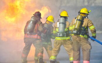 Four firefighters use a hose to put out a fire. One wears a red hard hat while the other wear yellow ones.