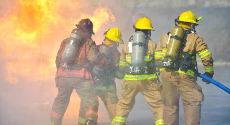 Four firefighters use a hose to put out a fire. One wears a red hard hat while the other wear yellow ones.