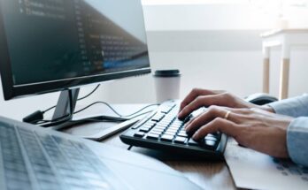 A man types on a keyboard. Next to him is another keyboard and monitor and a to-go coffee cup.