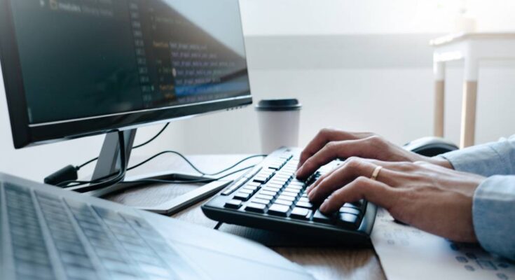 A man types on a keyboard. Next to him is another keyboard and monitor and a to-go coffee cup.