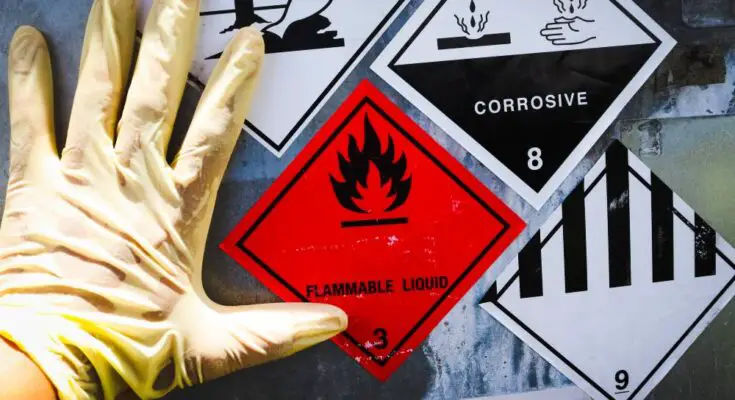 Close-up on a gloved hand touching a container that has warning stickers on it that say FLAMMABLE LIQUID and CORROSIVE.