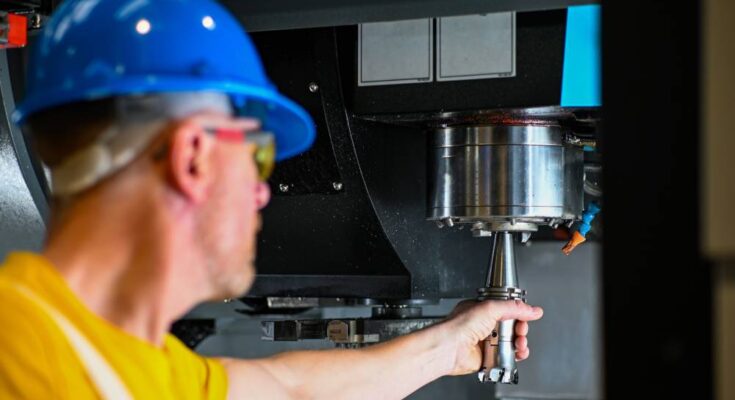 A man in a yellow shirt and blue hard hat reaching out with a tool to change the drill bit on a CNC machine.