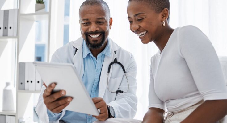 A woman in a white shirt and a male doctor in a lab coat sitting together in an office. They are looking at a tablet and smiling.