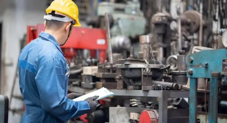 A worker wearing a blue work shirt and yellow hard hat holds a clipboard, looking over equipment at a factory.