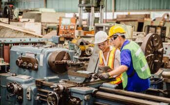 A man and woman on a factory floor wear bright vests and hardhats as they inspect a large machine with an electronic tablet.