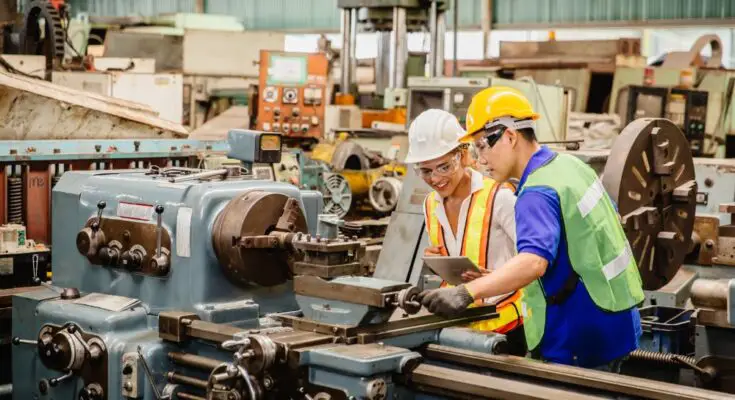 A man and woman on a factory floor wear bright vests and hardhats as they inspect a large machine with an electronic tablet.