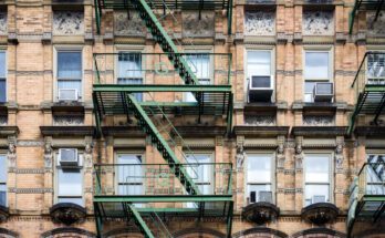 An old residential building with multiple stories and fire escapes. The building is brick and the escapes are green.