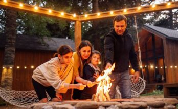 A father and mother are roasting hot dogs over an open fire pit with their young daughter and son in the backyard.