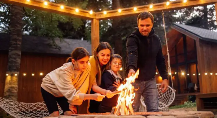 A father and mother are roasting hot dogs over an open fire pit with their young daughter and son in the backyard.