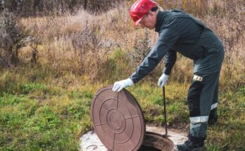 A worker wearing a green suit and a red helmet lifts the lid of a manhole cover to service a septic system.