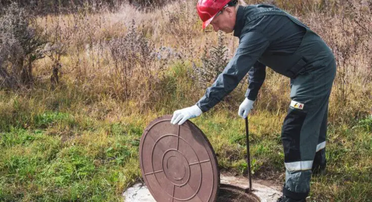 A worker wearing a green suit and a red helmet lifts the lid of a manhole cover to service a septic system.