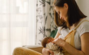 A woman in yellow overalls and a gray shirt sits on a tan couch, breastfeeding a baby in a yellow-and-white striped outfit.