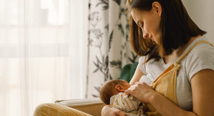 A woman in yellow overalls and a gray shirt sits on a tan couch, breastfeeding a baby in a yellow-and-white striped outfit.