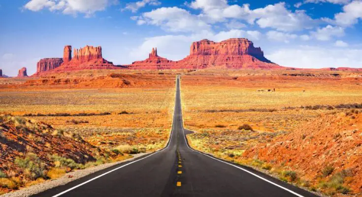 A paved road going through the center of Monument Valley toward distant mesas and buttes with a rocky landscape on each side.