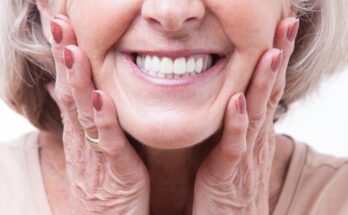 A close-up of an older woman with gray hair smiling widely and showing her teeth. Her hands are on the sides of her face.