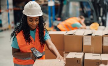 Female worker wearing a safety vest and helmet using a barcode scanner on a pile of cardboard boxes.