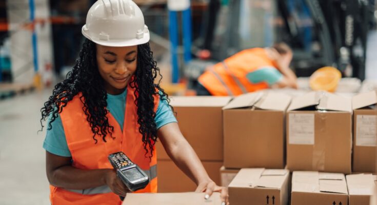 Female worker wearing a safety vest and helmet using a barcode scanner on a pile of cardboard boxes.