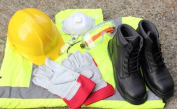 A collection of personal protective equipment including a hi-vis vest, hard hat, and steel-toed boots sit on the floor.