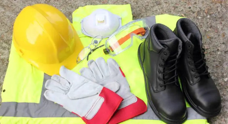 A collection of personal protective equipment including a hi-vis vest, hard hat, and steel-toed boots sit on the floor.