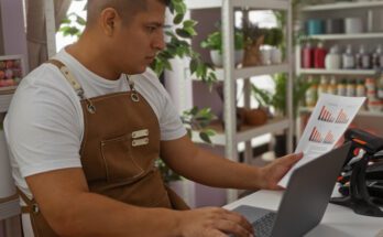 A man in a white shirt and brown apron sits at a desk in front of a silver laptop, holding a piece of paper with graphs on it.