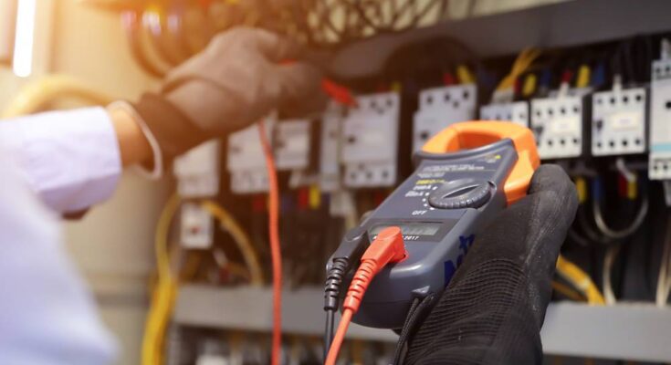 An engineer using a digital multimeter checks the electrical current voltage at a circuit breaker board.