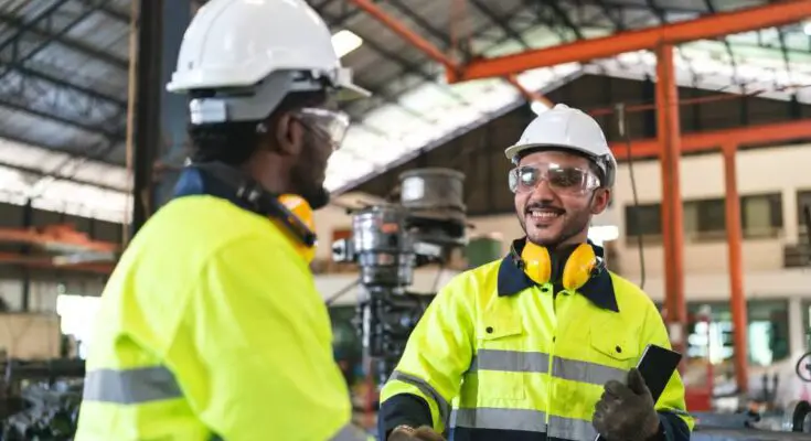 Two men wearing hard hats, safety glasses, and hi-vis jackets shake hands while standing in a manufacturing facility.